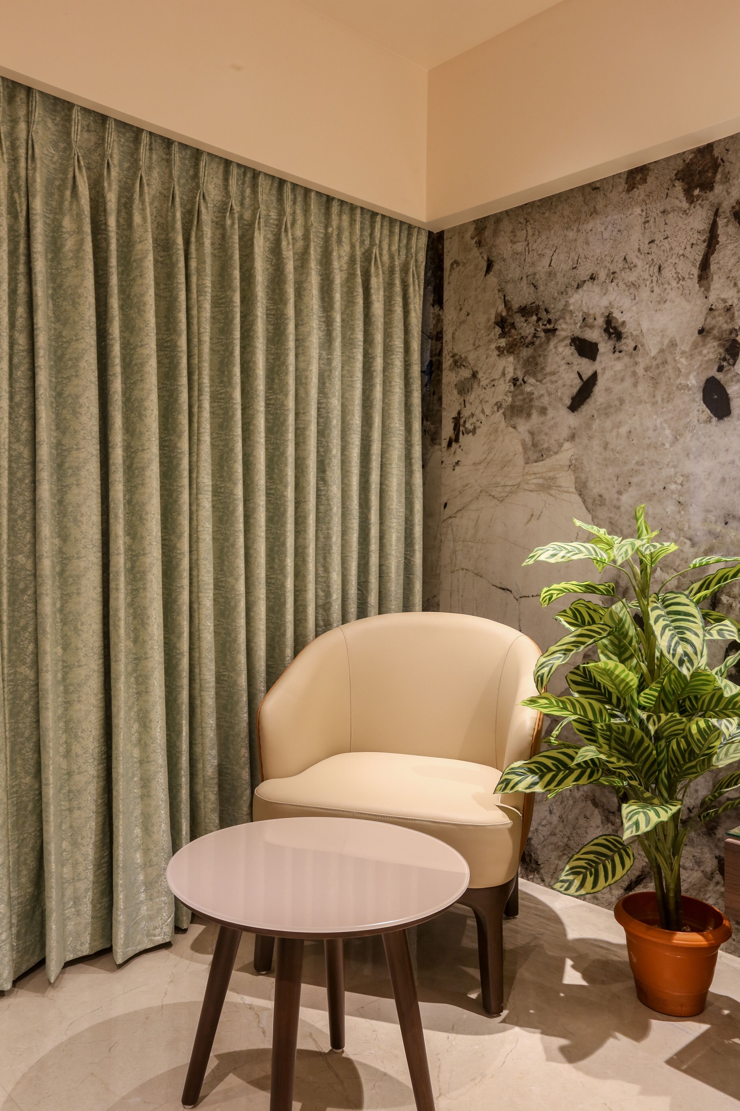 Una habitación con un sillón color beige, una mesa redonda pequeña, una planta en maceta y una cortina.
