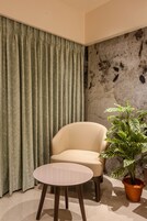 Una habitación con un sillón color beige, una mesa redonda pequeña, una planta en maceta y una cortina.