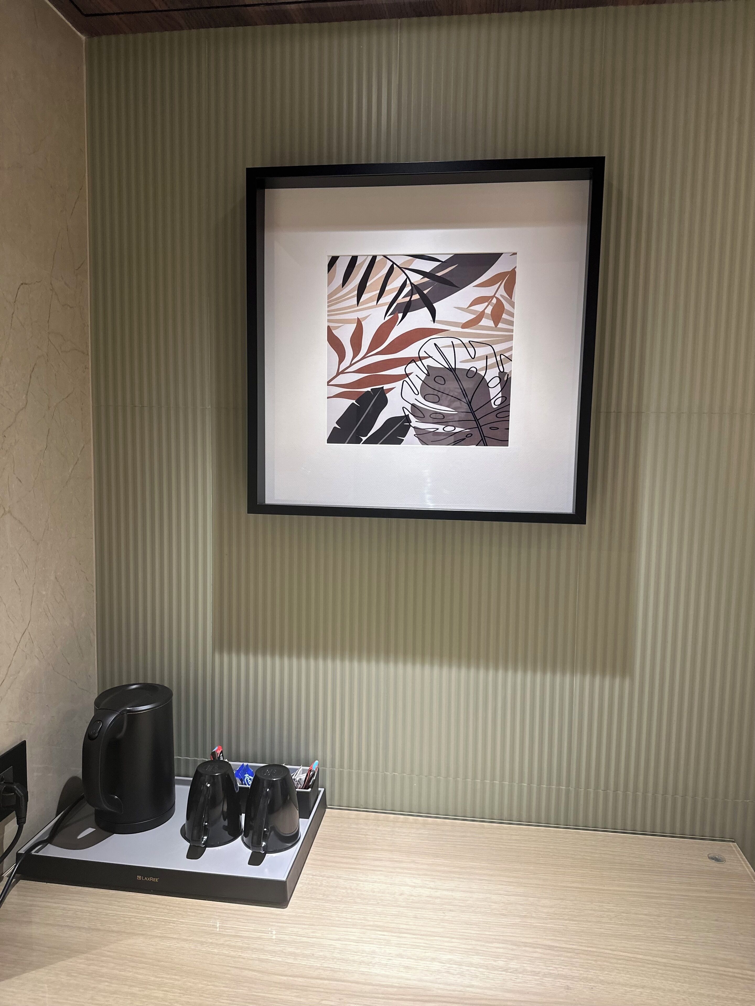 A framed artwork of leaves on a wall, a black coffee maker on a table, and a LEXREX brand kettle.