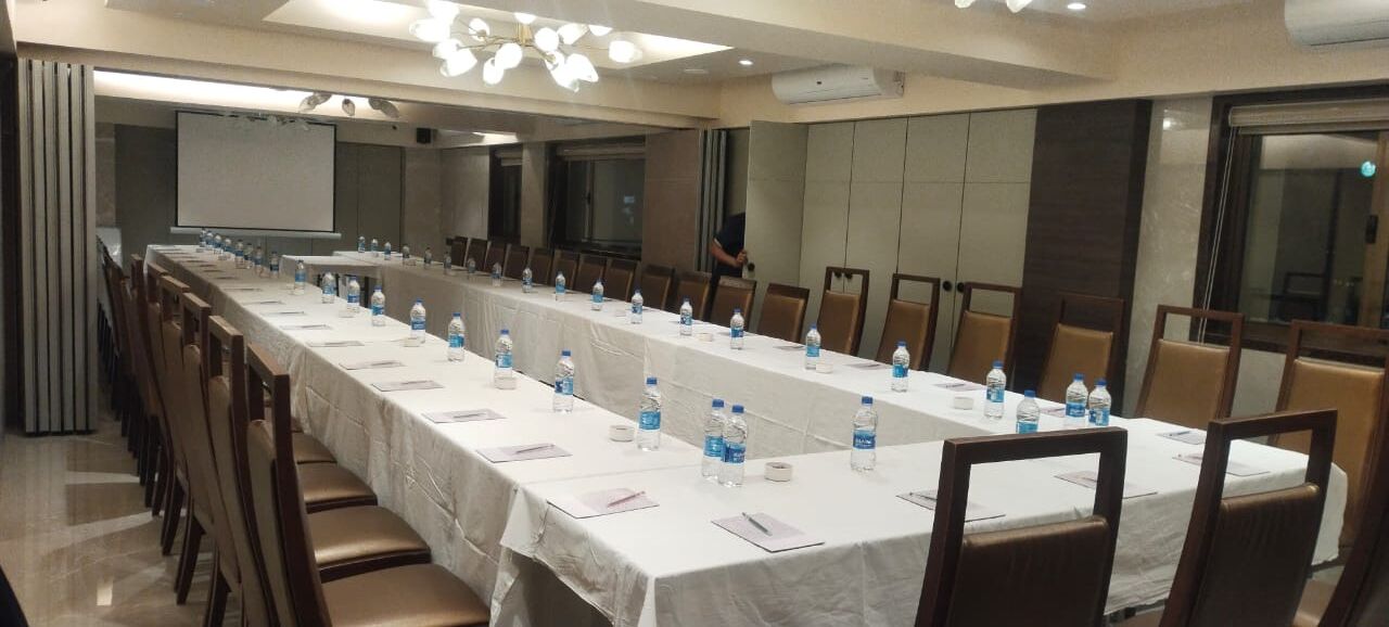 Un salón de conferencias con una larga mesa, sillas, botellas de agua y una pantalla para proyector.