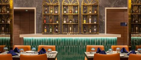 Zona de bar con tres estantes llenos de botellas de licores, asientos anaranjados y una barra verde.