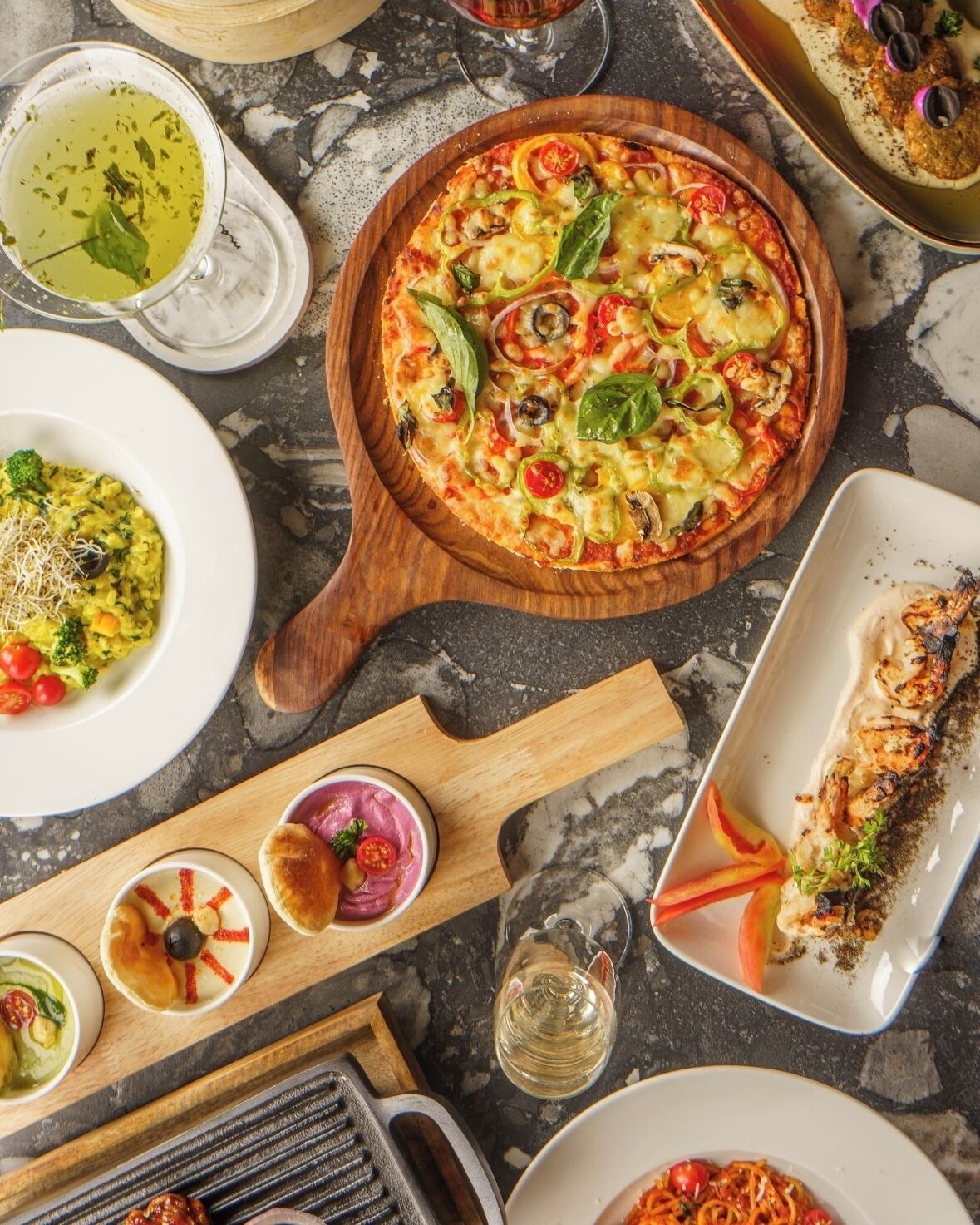 A variety of dishes including a pizza with vegetables, a bowl of soup, and a plate of grilled fish, all arranged on a marble table.