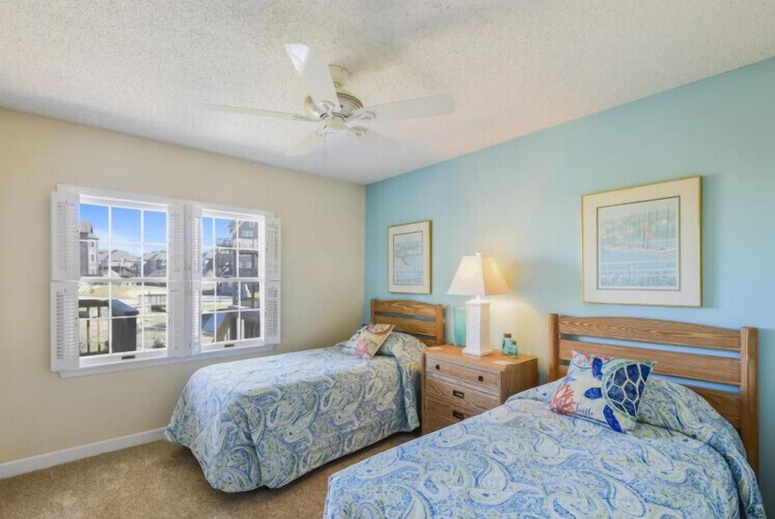 Un dormitorio con dos camas, ventilador de techo, ventana con contraventanas y cuadros en la pared.