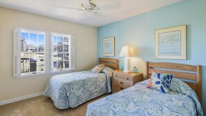 Une chambre avec deux lits, un ventilateur de plafond, une fenêtre avec des volets et des œuvres d’art encadrées accrochées au mur.