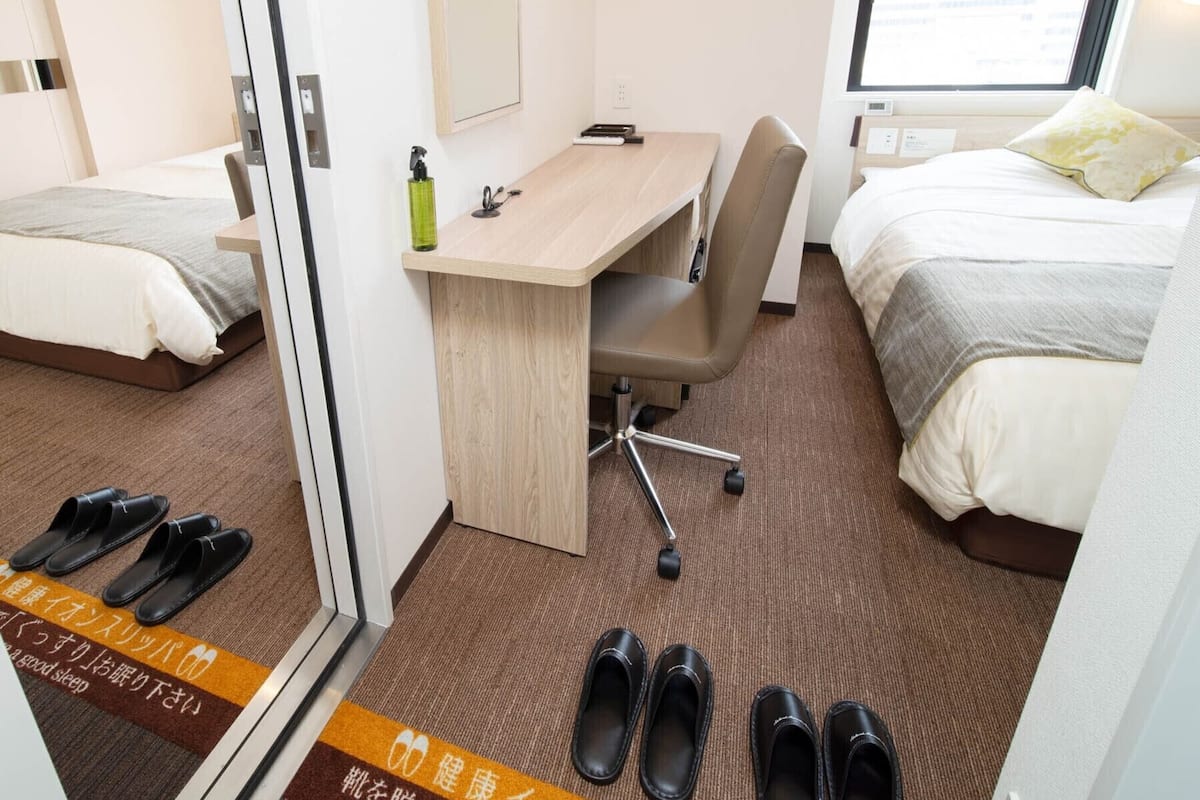 A hotel room with a desk, two beds, a window, and slippers on the floor.