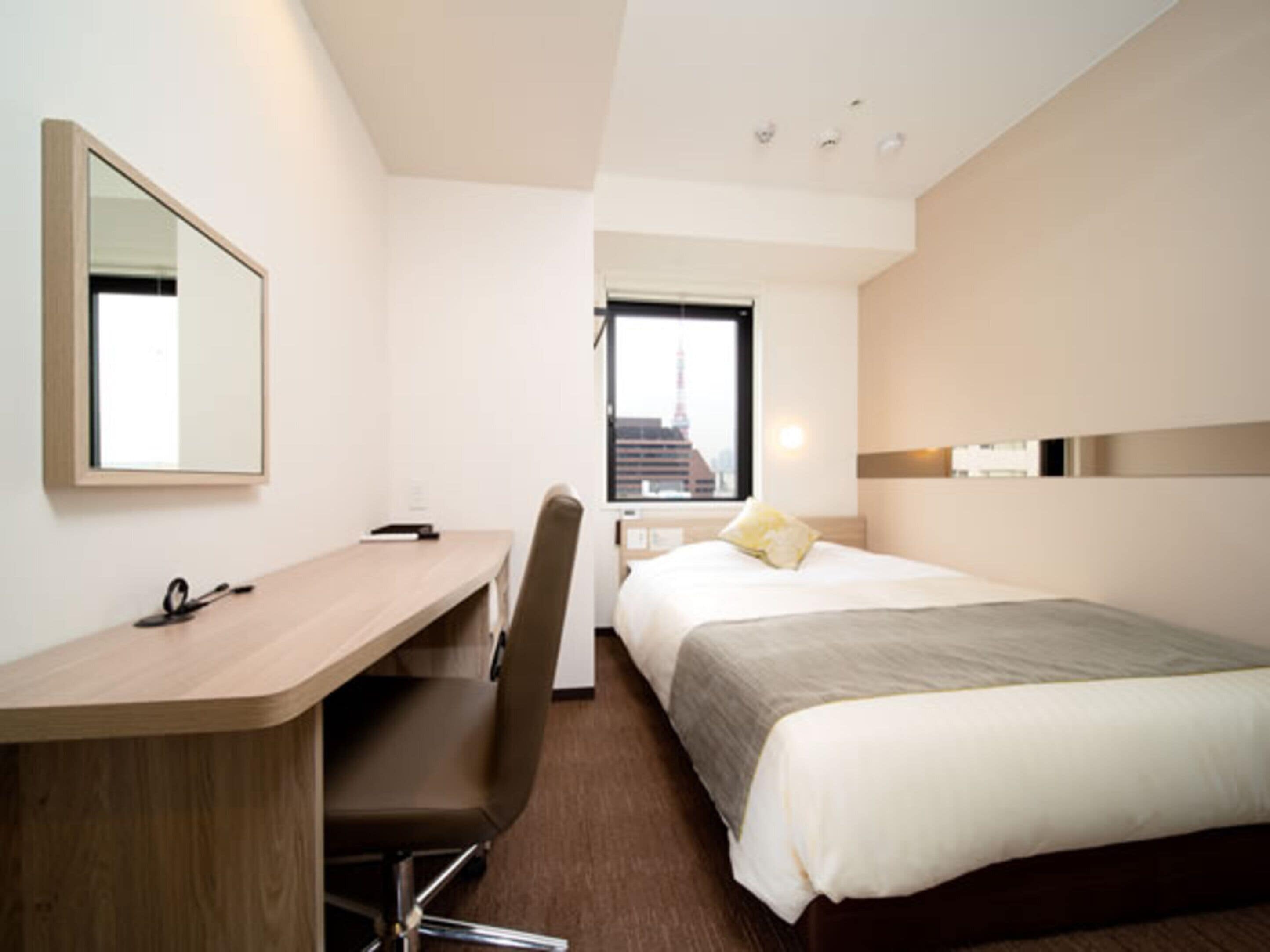A hotel room with a bed, a desk, a chair, a mirror, and a window with a city view.