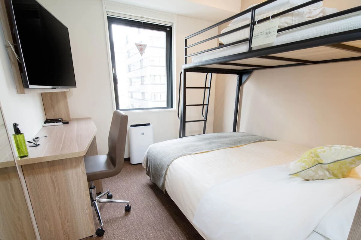A hotel room with a bunk bed, a desk with a chair, a TV, and a window with a view of buildings.