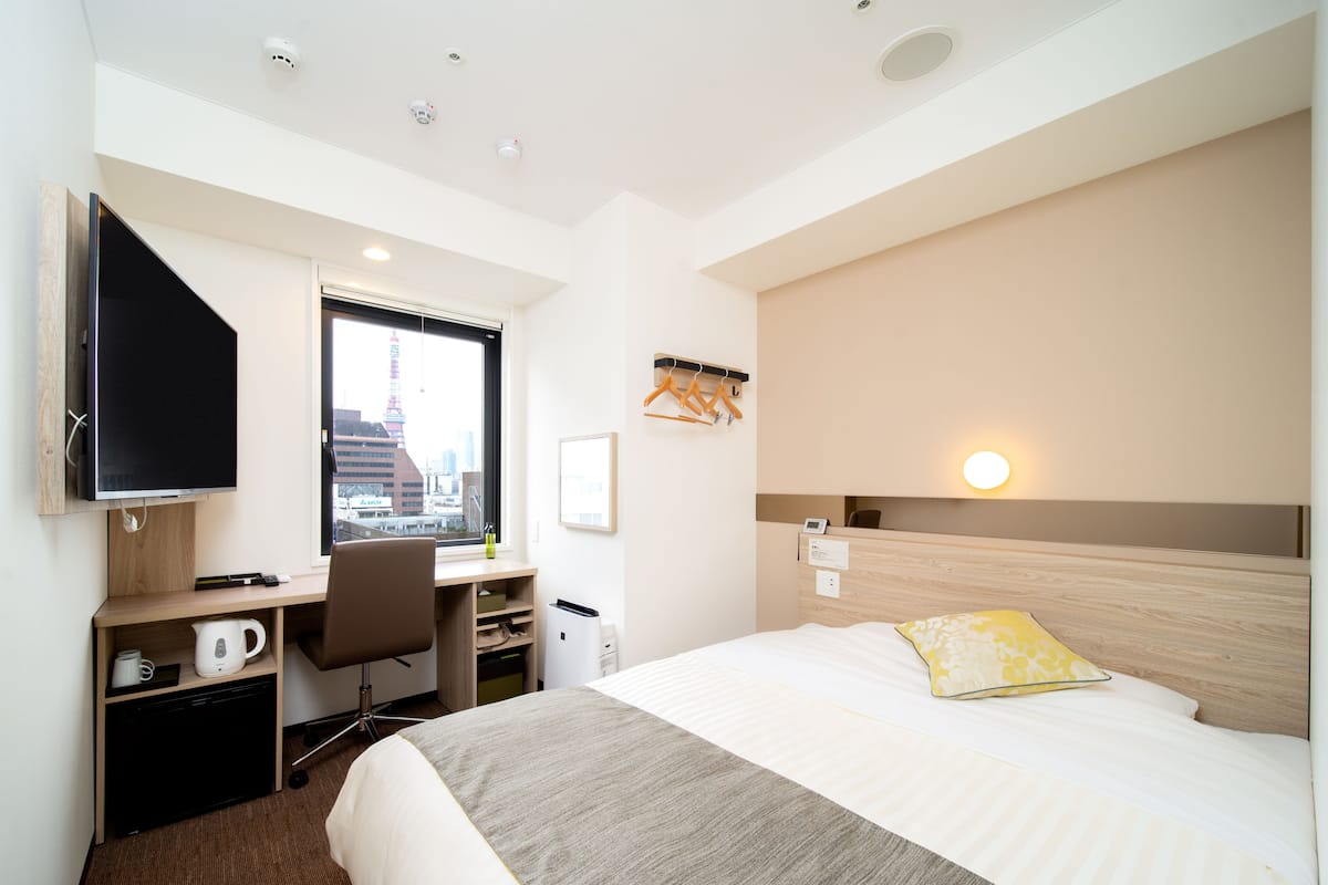 A hotel room with a bed, a desk, a television, a window with a city view, and a bedside table with a lamp.