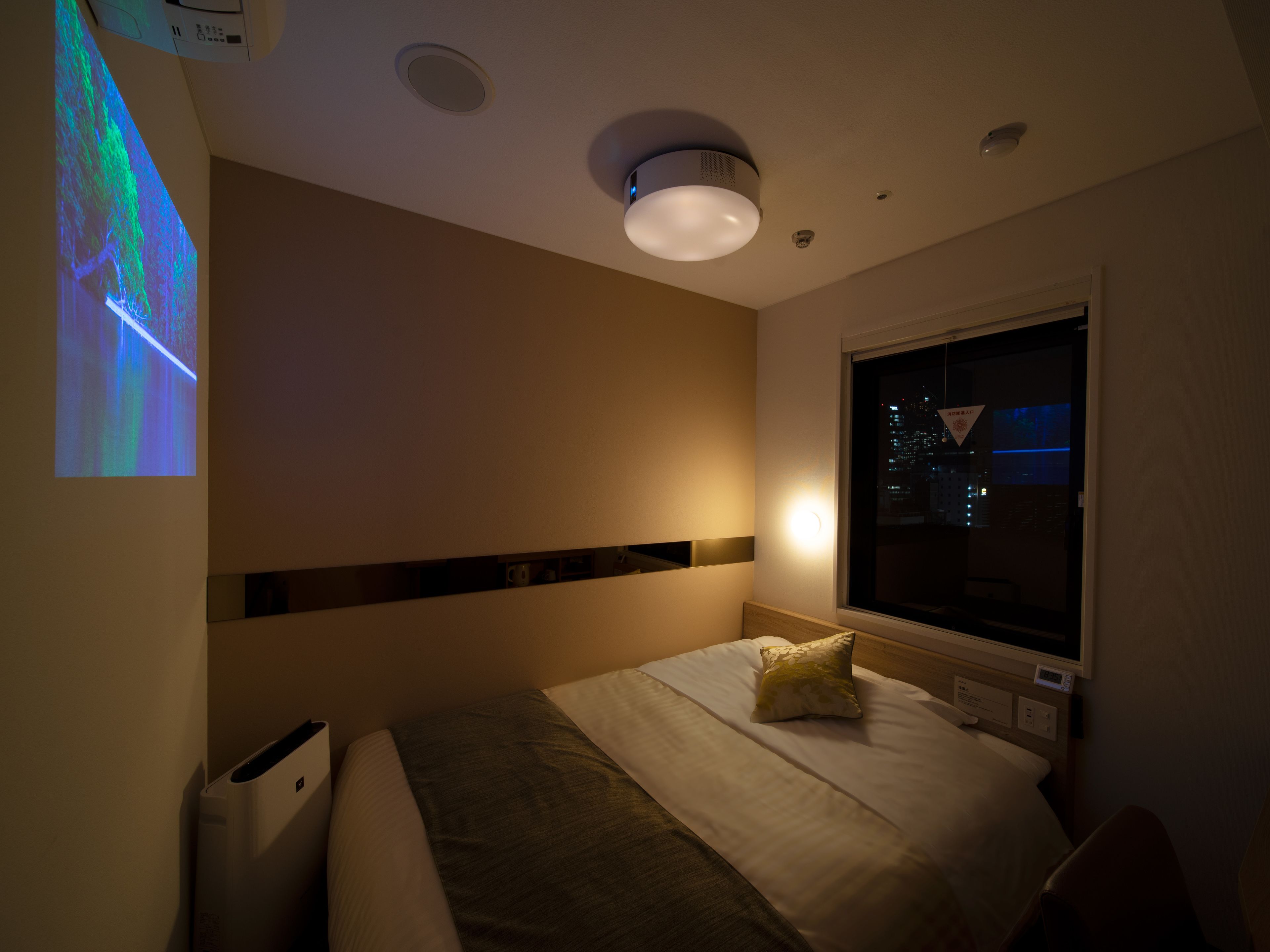 A hotel room with a bed, a window, a wall-mounted projector, and a ceiling light.