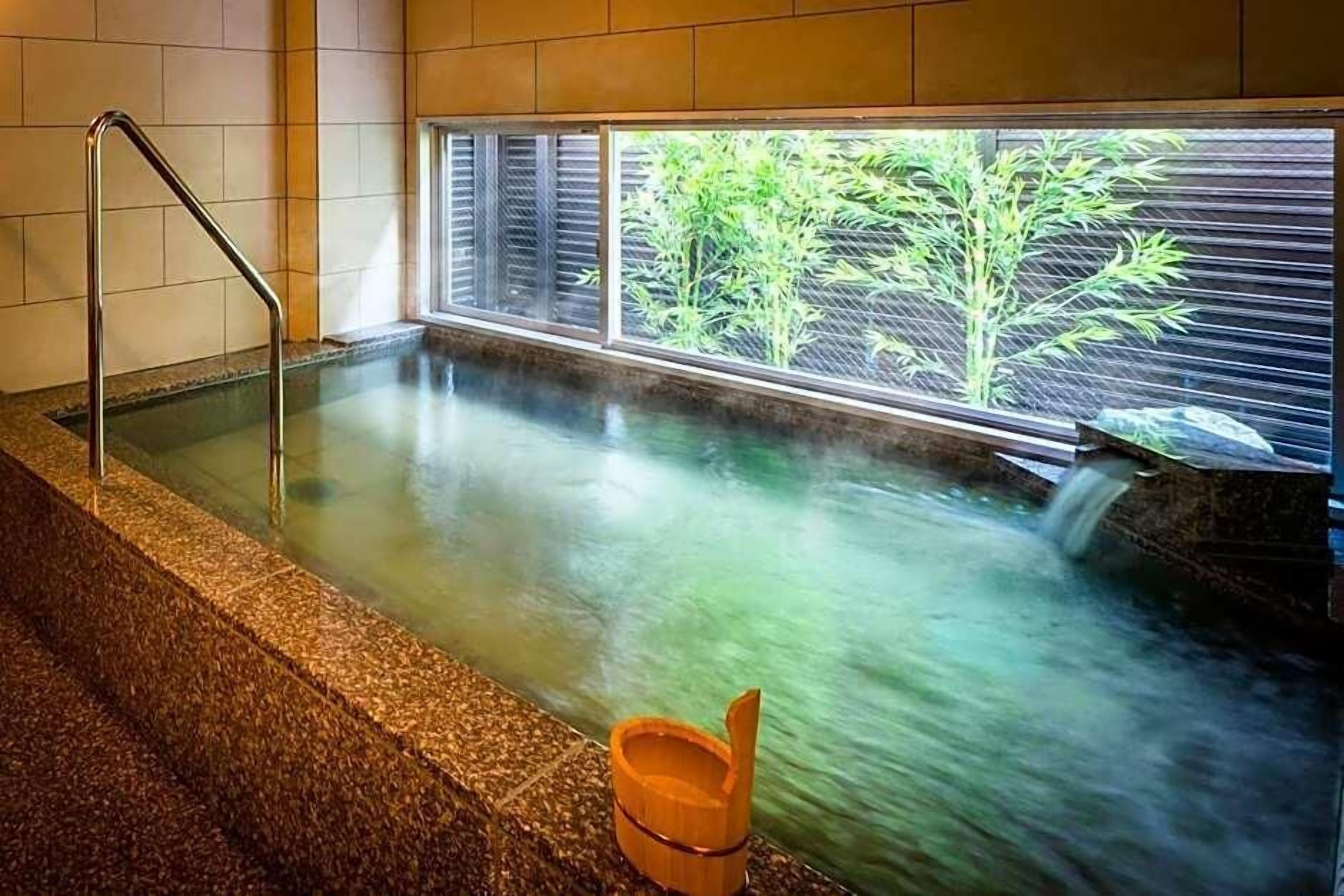 A hot spring bath with a view of bamboo outside, a metal ladder, and a wooden bucket.
