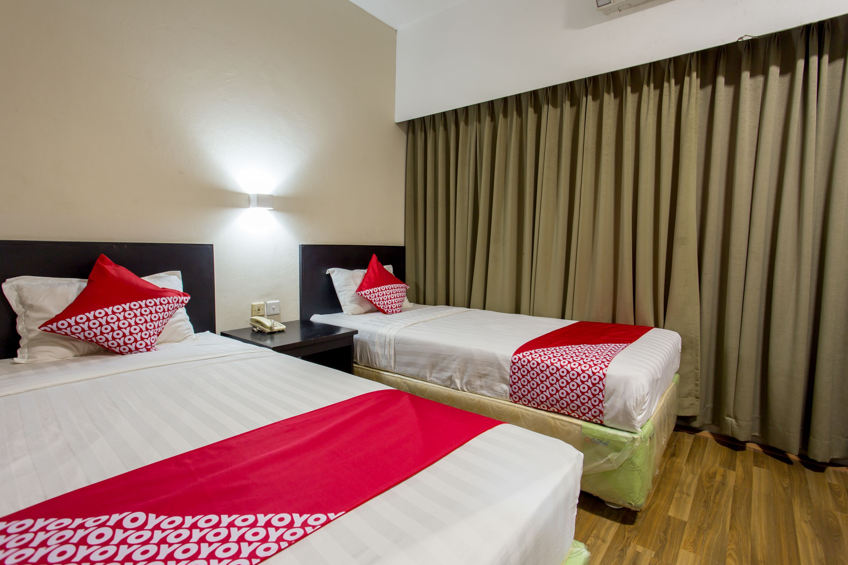 Quarto de hotel com duas camas, cada uma com colcha de padrão vermelho e branco, uma mesa de cabeceira com telefone e uma luminária fixada na parede.