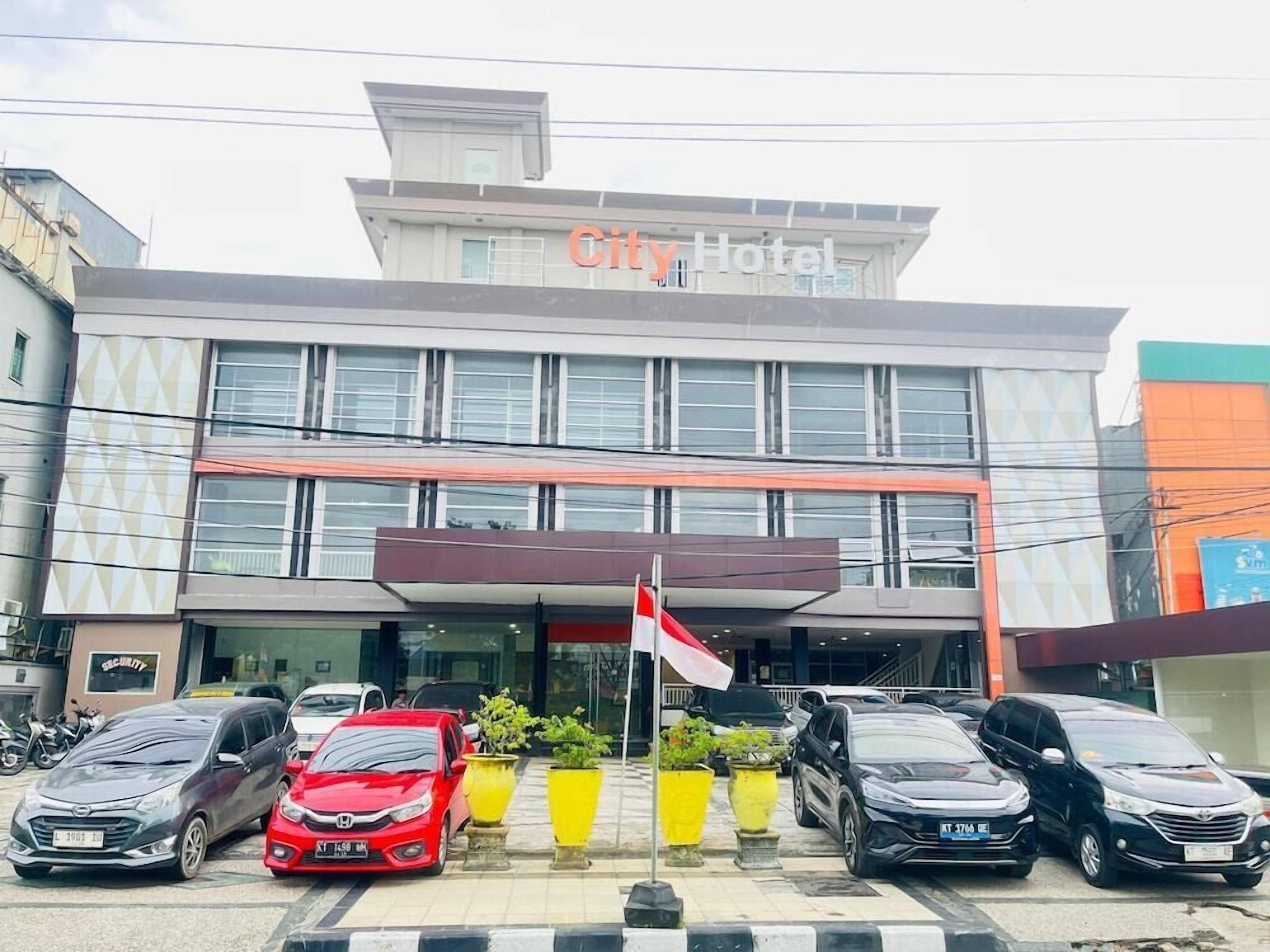 Een hotel met een moderne gevel, meerdere auto's geparkeerd ervoor en een vlaggenmast met een vlag.