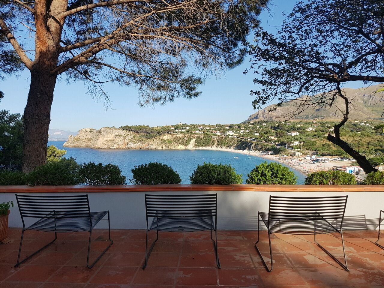 A terrace with three metal chairs and a view of a coastal landscape with cliffs and a beach.