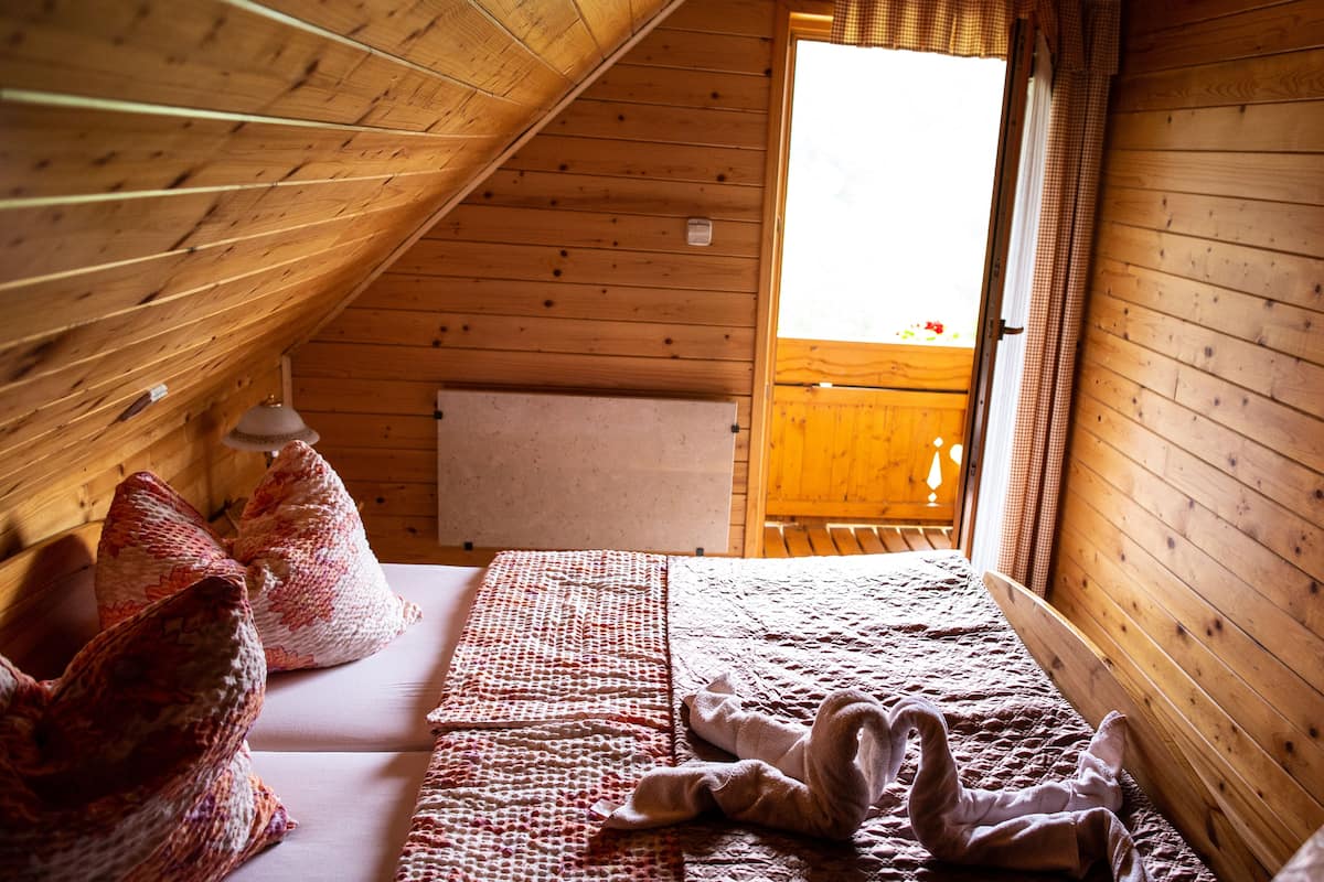 Une chambre mansardée chaleureuse, avec un plafond en pente, un lit recouvert d’une literie à motifs et une fenêtre agrémentée de rideaux.