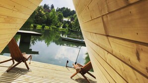 Eine Holzterrasse mit zwei Stühlen, die auf einen ruhigen See blicken, umgeben von viel Grün und blauem Himmel.