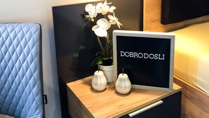 Ein ordentlich eingerichtetes Hotelzimmer mit einem Bett, einem Nachttisch mit einer Lampe, einer Topforchidee und einem Schild mit der Aufschrift 'DOBRODOSLI'.