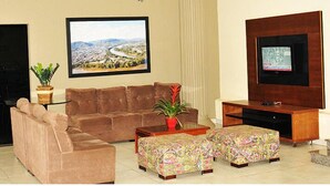 A living room with a brown sofa, a coffee table, two patterned ottomans, a TV on a wooden stand, and a framed cityscape picture on the wall. - Hotel Boa Viagem (Barra do Garcas)