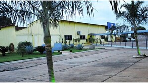 A building with a yellow facade, a covered outdoor area, and a paved courtyard with palm trees. - Hotel Boa Viagem (Barra do Garcas)