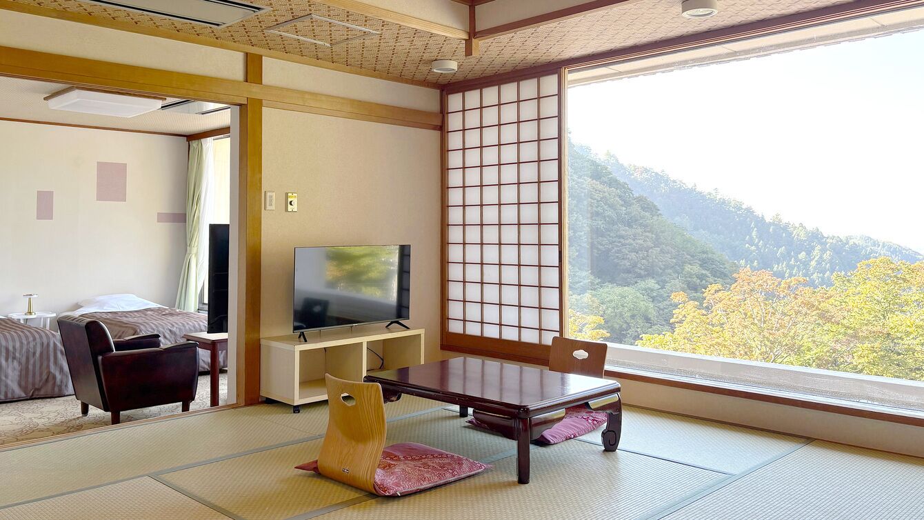 Een traditionele Japanse kamer met tatami vloeren, een televisie, een bank en een schuifdeur met uitzicht op een bebost glooiend landschap.