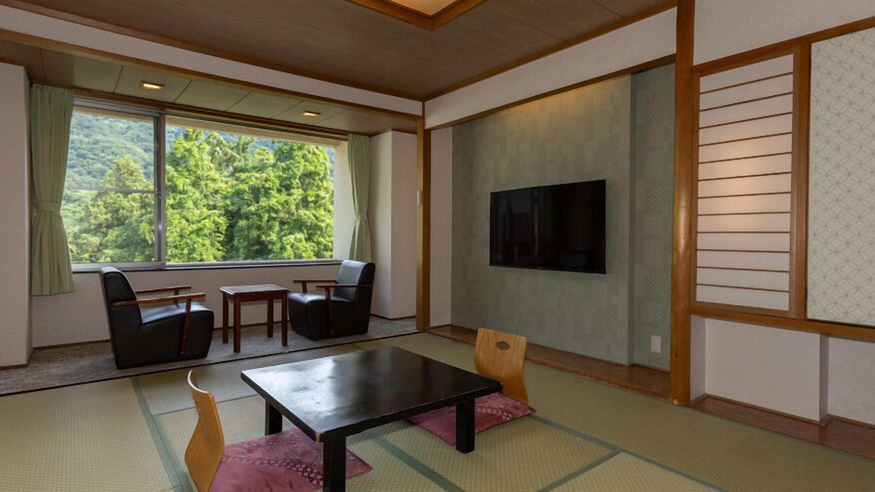 Een traditionele Japanse kamer met tatami vloerbedekking, een lage tafel, stoelen, een televisie en een groot raam met uitzicht op bomen.