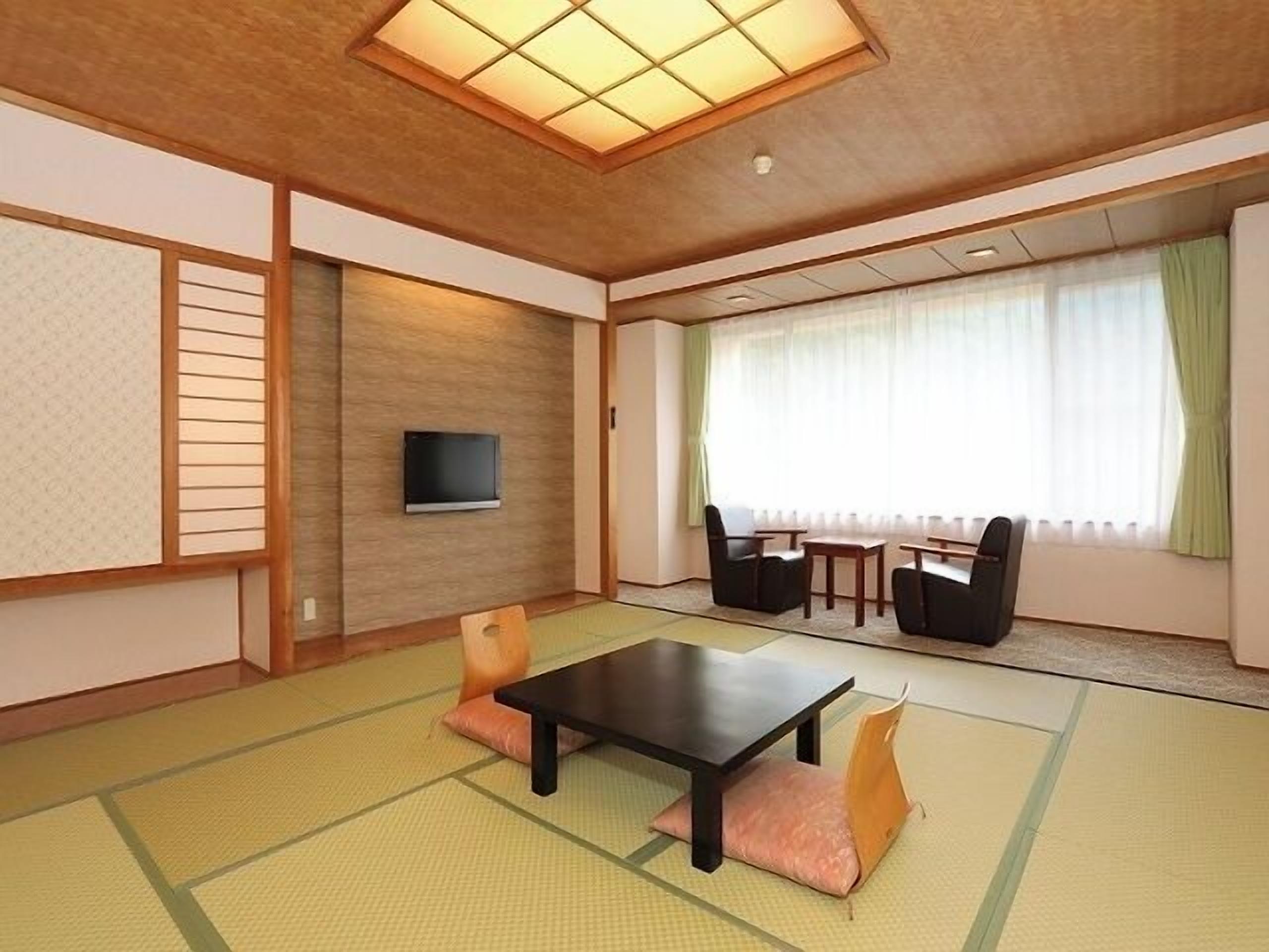 Una habitación japonesa tradicional con suelo de tatami, una mesa baja, sillas, un televisor y un amplio ventanal con cortinas.