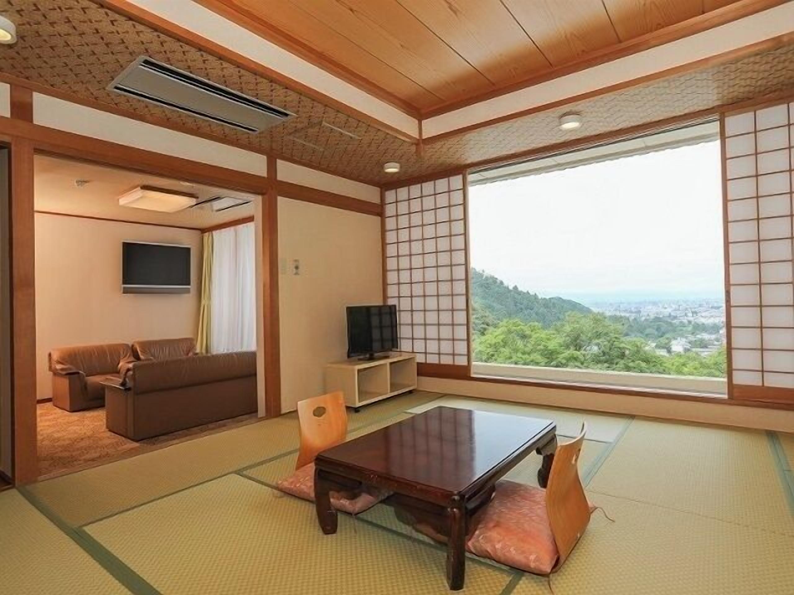 Una habitación japonesa tradicional con suelo de tatami, una mesa de madera baja, un sofá, un televisor y un amplio ventanal con vistas a un paisaje urbano.