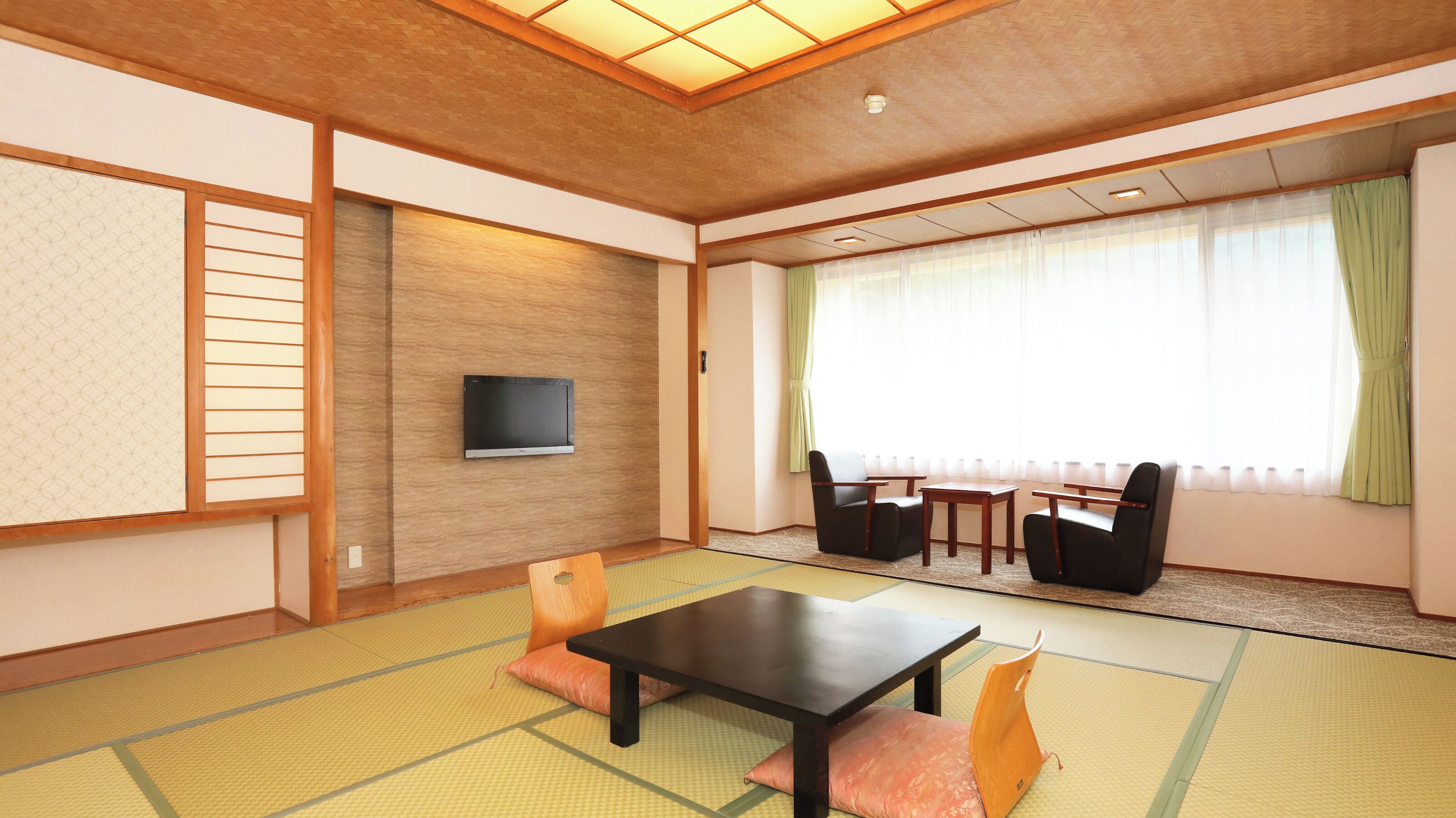 Una habitación de estilo japonés tradicional con suelo de tatami, televisor montado en la pared y zona de estar con una mesa baja y sillas.