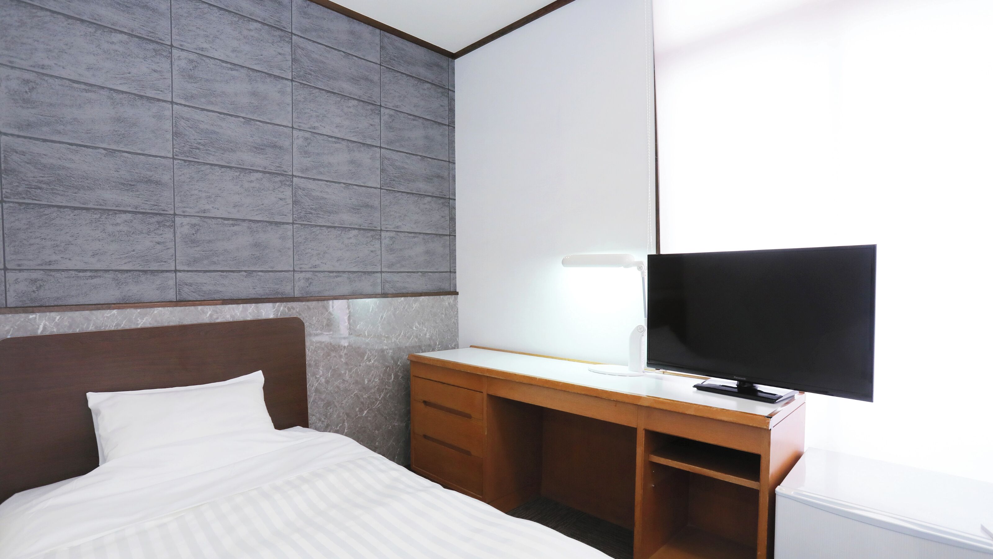 Habitación de hotel con cama, escritorio y televisor de pantalla plana, y pared con azulejos grises.