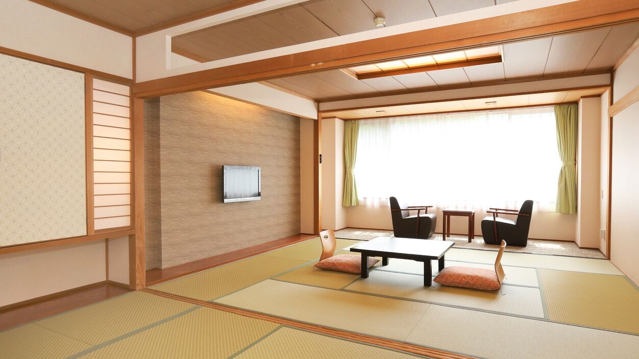 Een traditionele Japanse kamer met tatami vloerbedekking, een lage tafel, stoelen, een televisie en uitzicht buiten door een raam met gordijnen.
