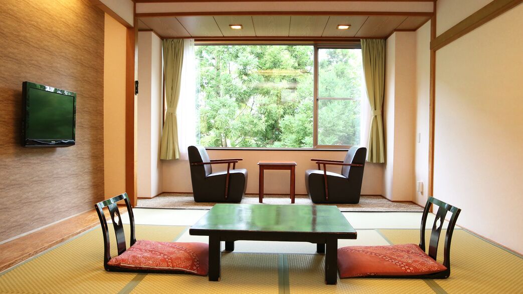 Een kamer met tatami vloeren, een salontje met twee stoelen, een televisie aan de muur, en een groot raam met gordijnen.