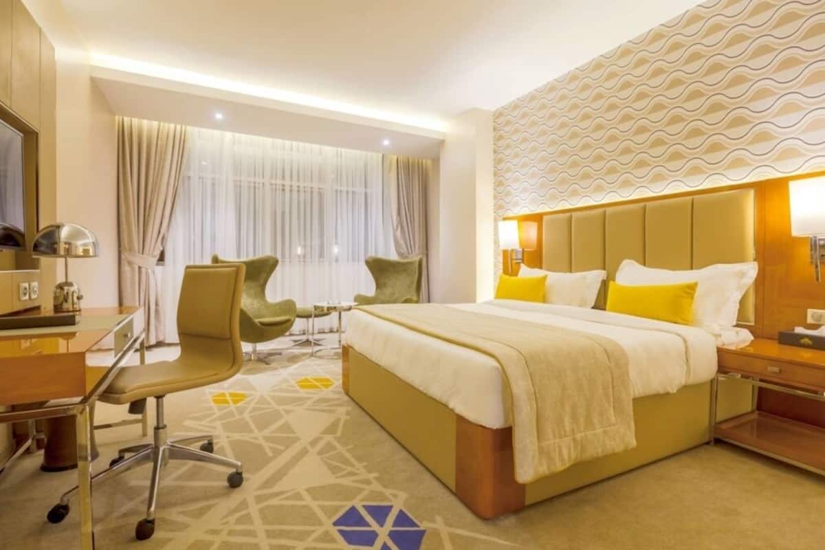 A hotel room with a bed, a desk with a lamp, a chair, and a window with curtains.