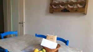 A dining table with a wooden board holding lemons and a paper, blue chairs around it, and a wooden shelf above with plates.