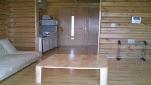 A log cabin interior with a wooden floor, a white sofa, a wooden coffee table, a kitchen area, and a door leading to another room.