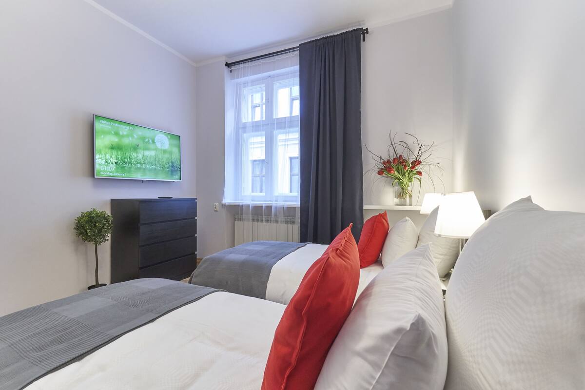 A hotel room with two beds, a flat-screen TV mounted on the wall, a window with curtains, a bedside table with a lamp, and a small potted plant.