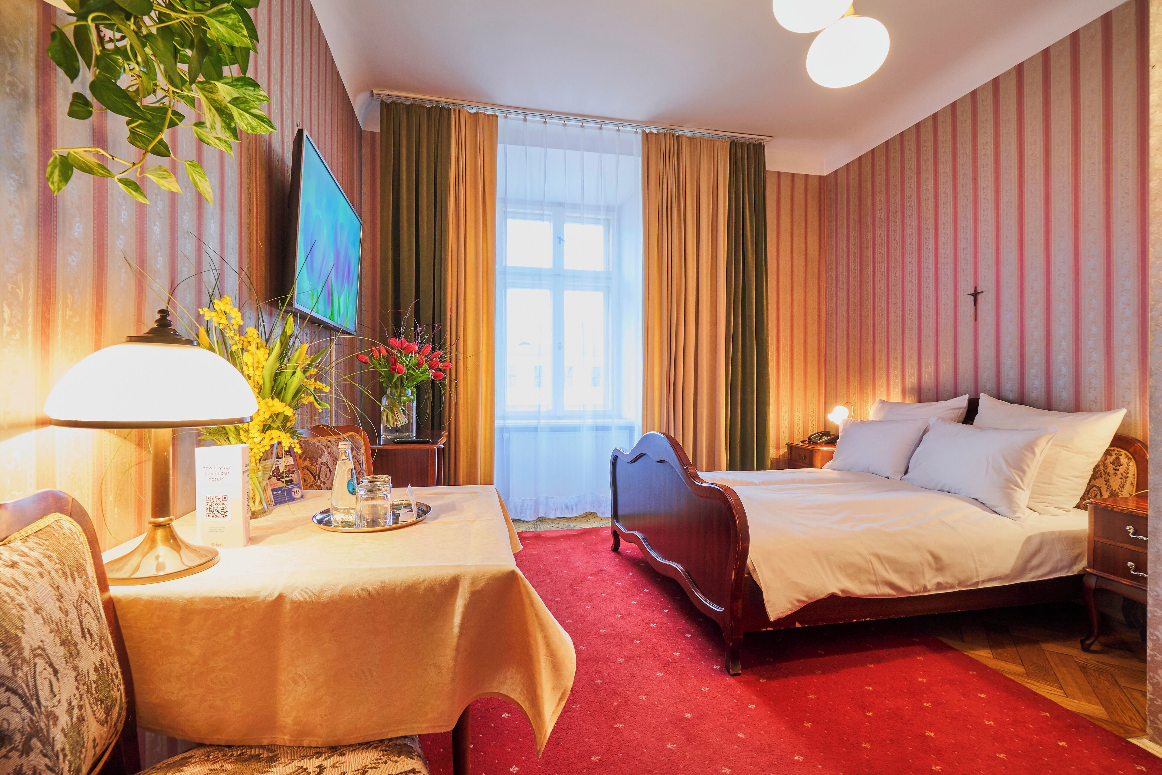 A hotel room with a bed, a bedside table with a lamp, a small table with a vase of flowers, a TV mounted on the wall, and a window with curtains.