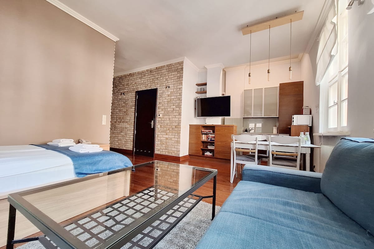 A modern hotel room with a sofa, a glass-top table, a TV, a kitchen area, and a brick wall.