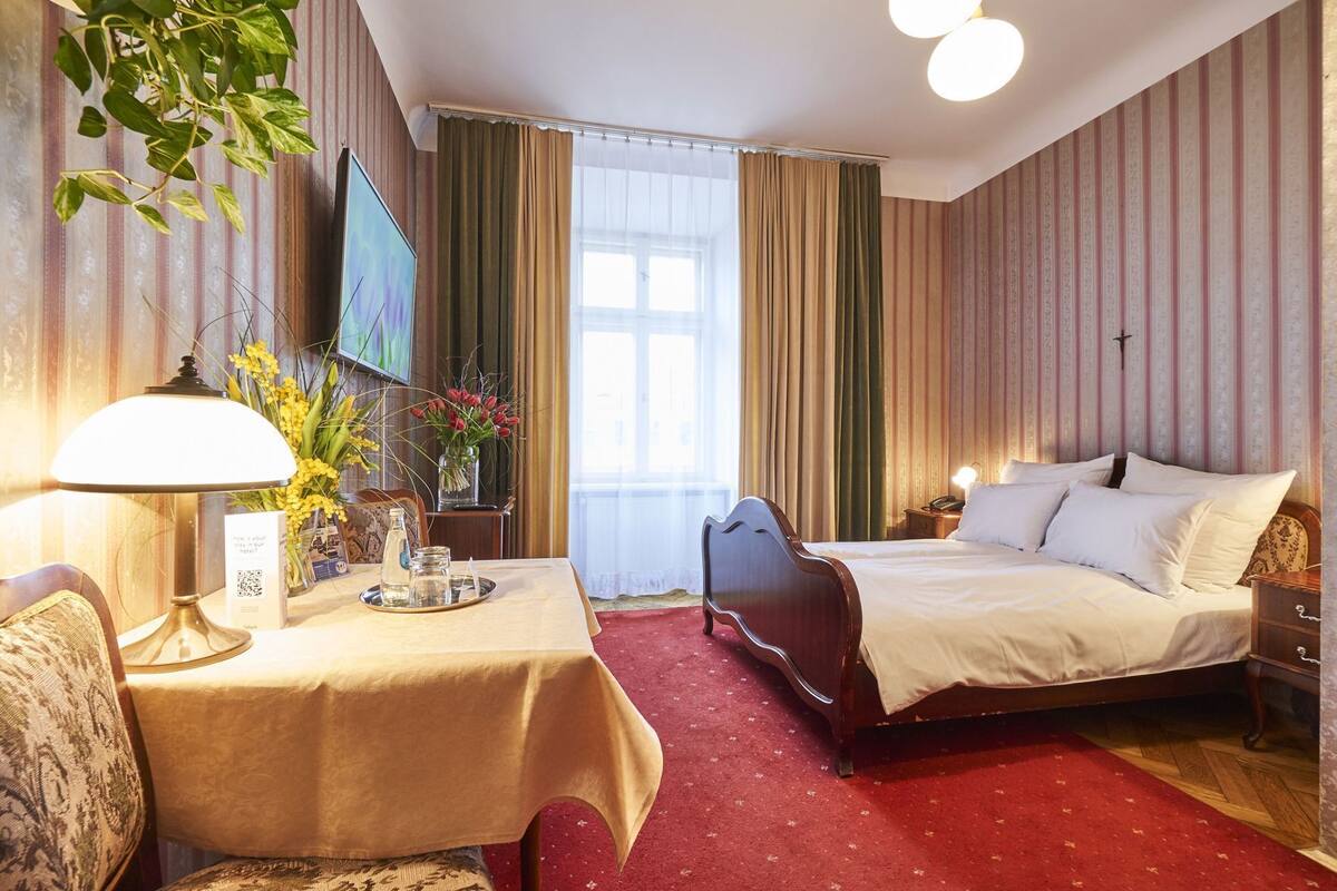 A hotel room with a bed, a desk with a lamp, a TV, and a window with curtains.