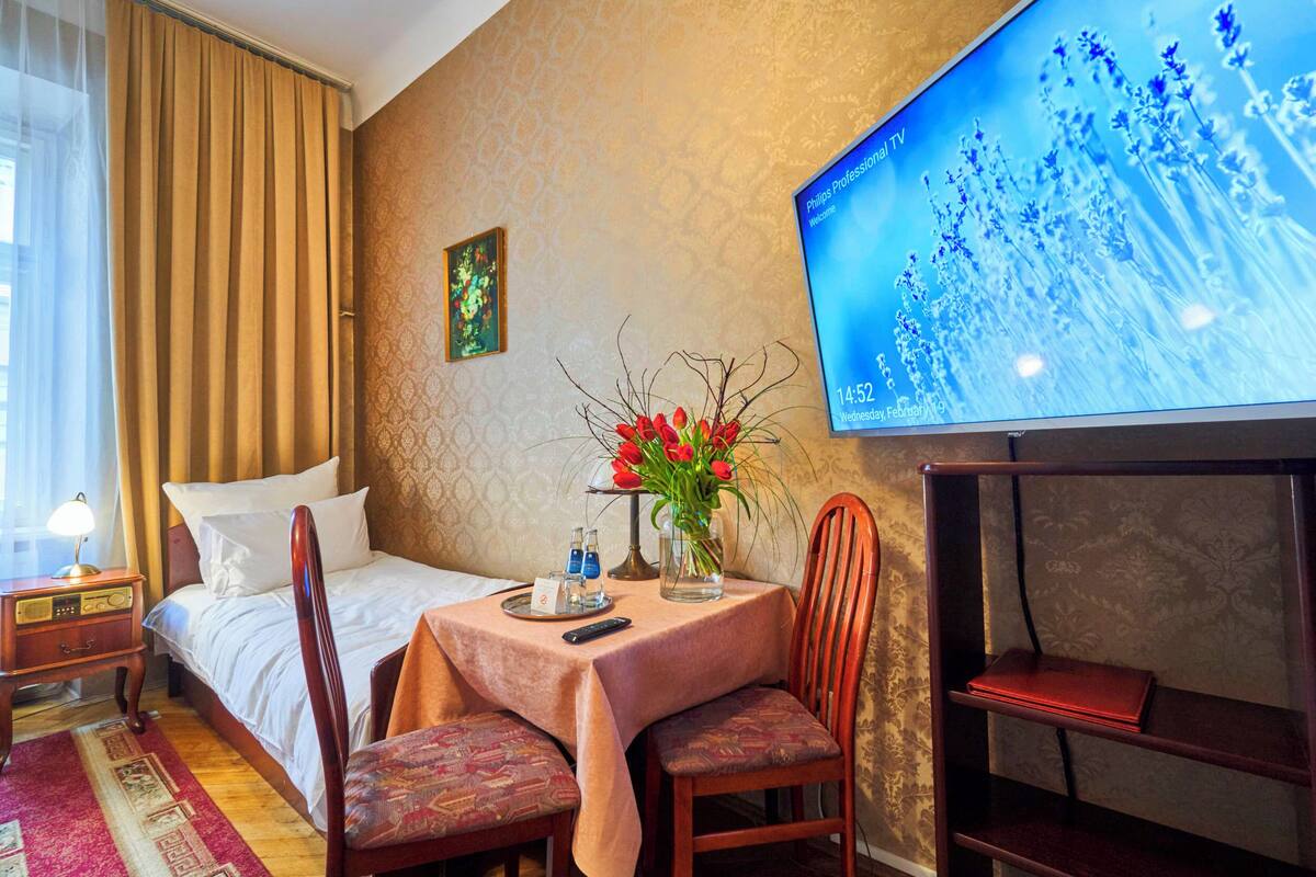 A hotel room with a bed, a desk with a lamp, a TV, a table with a vase of flowers, and a chair.