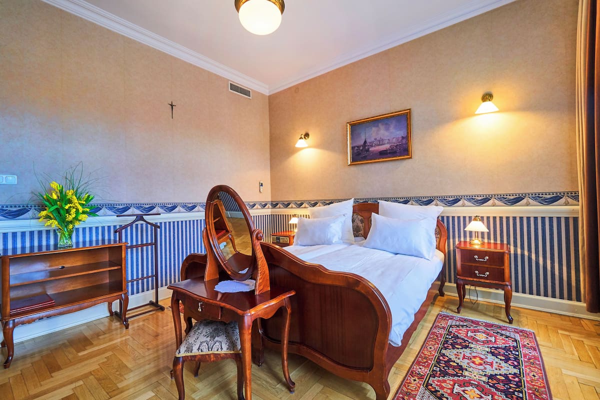 A hotel room with a large bed, a desk with a chair, a small table, a mirror, a nightstand, and a wall-mounted lamp.