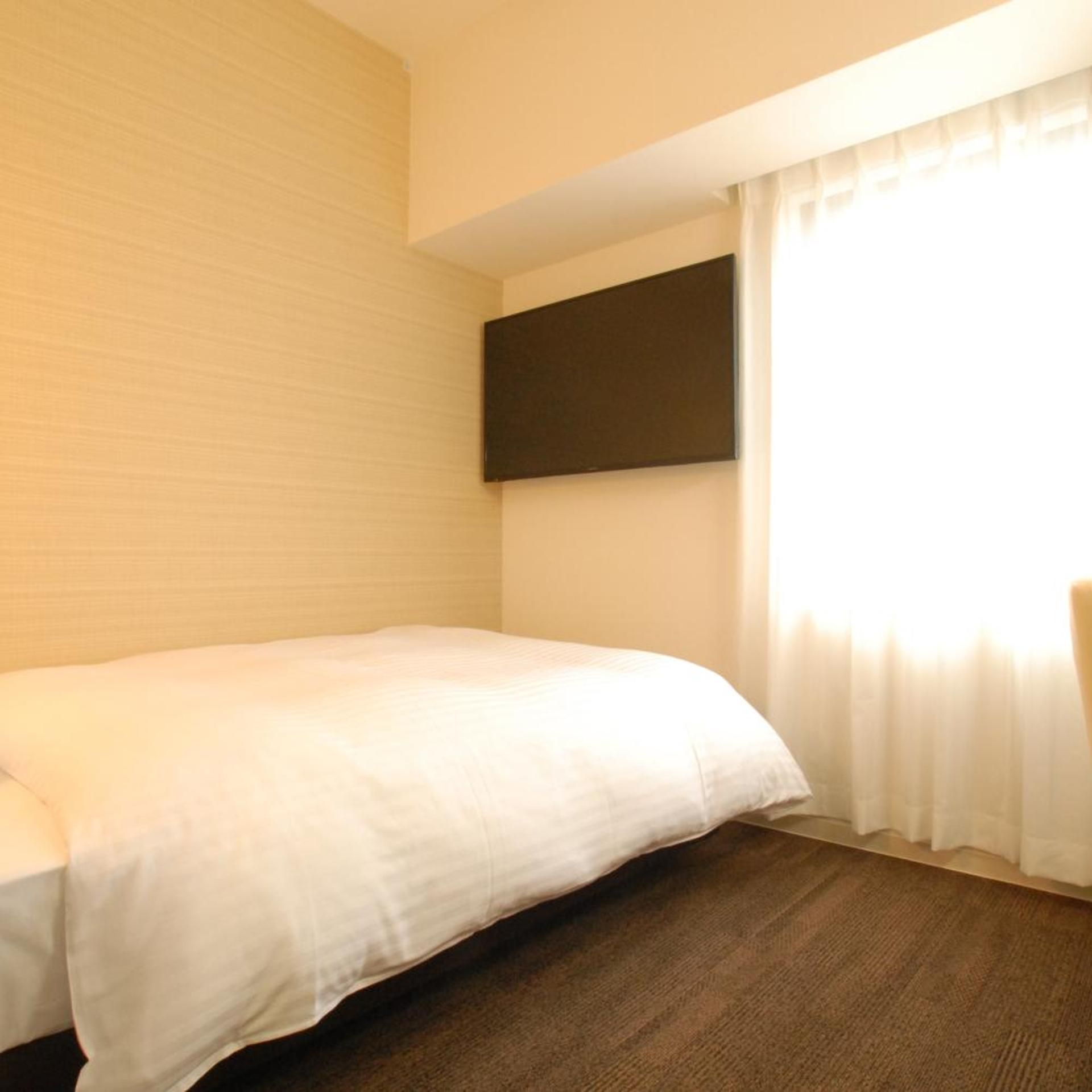A hotel room with a bed, a flat-screen TV mounted on the wall, and a window with curtains.