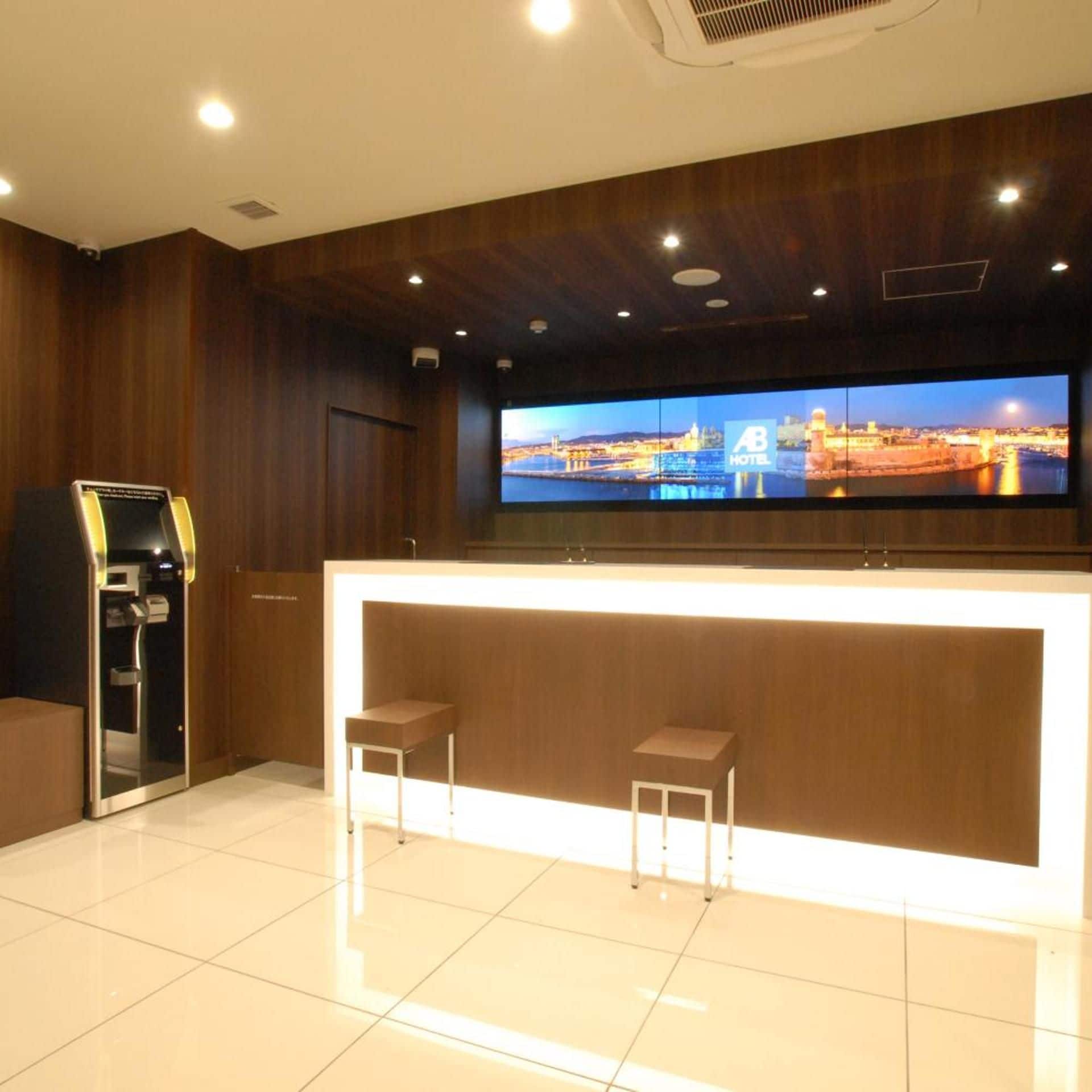 A hotel reception area with a modern design, featuring a reception desk, two bar stools, a large flat-screen TV displaying a cityscape, and a self-service kiosk.