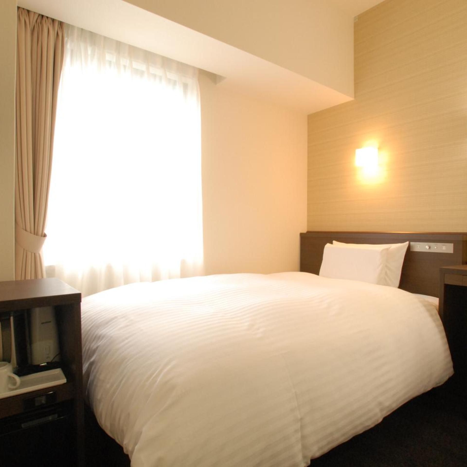 Habitación de hotel con una cama grande, una mesita de noche, una lámpara y una ventana con cortinas.