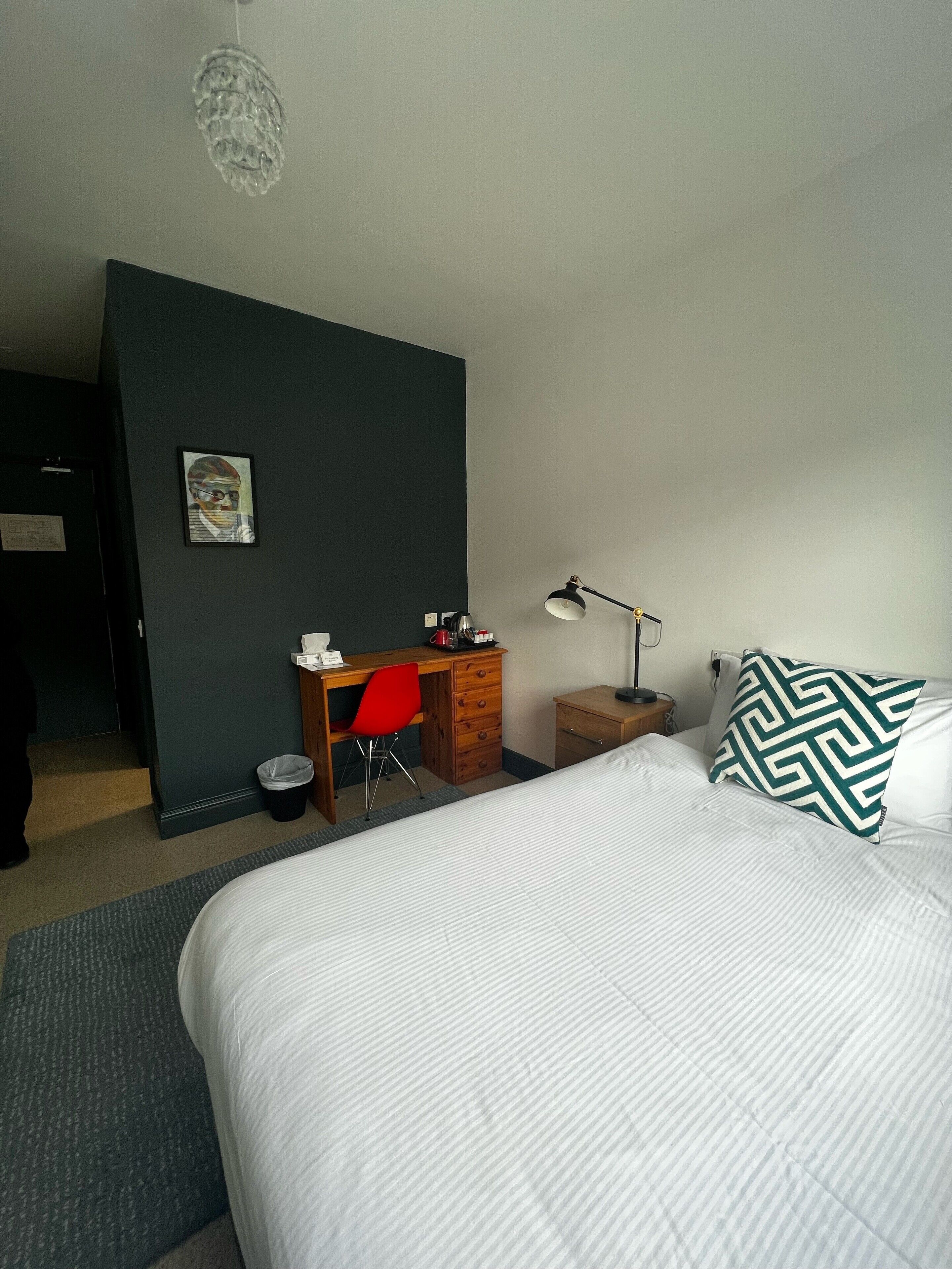 A hotel room with a bed, a desk with a chair, a lamp, a small cabinet, and a picture on the wall.
