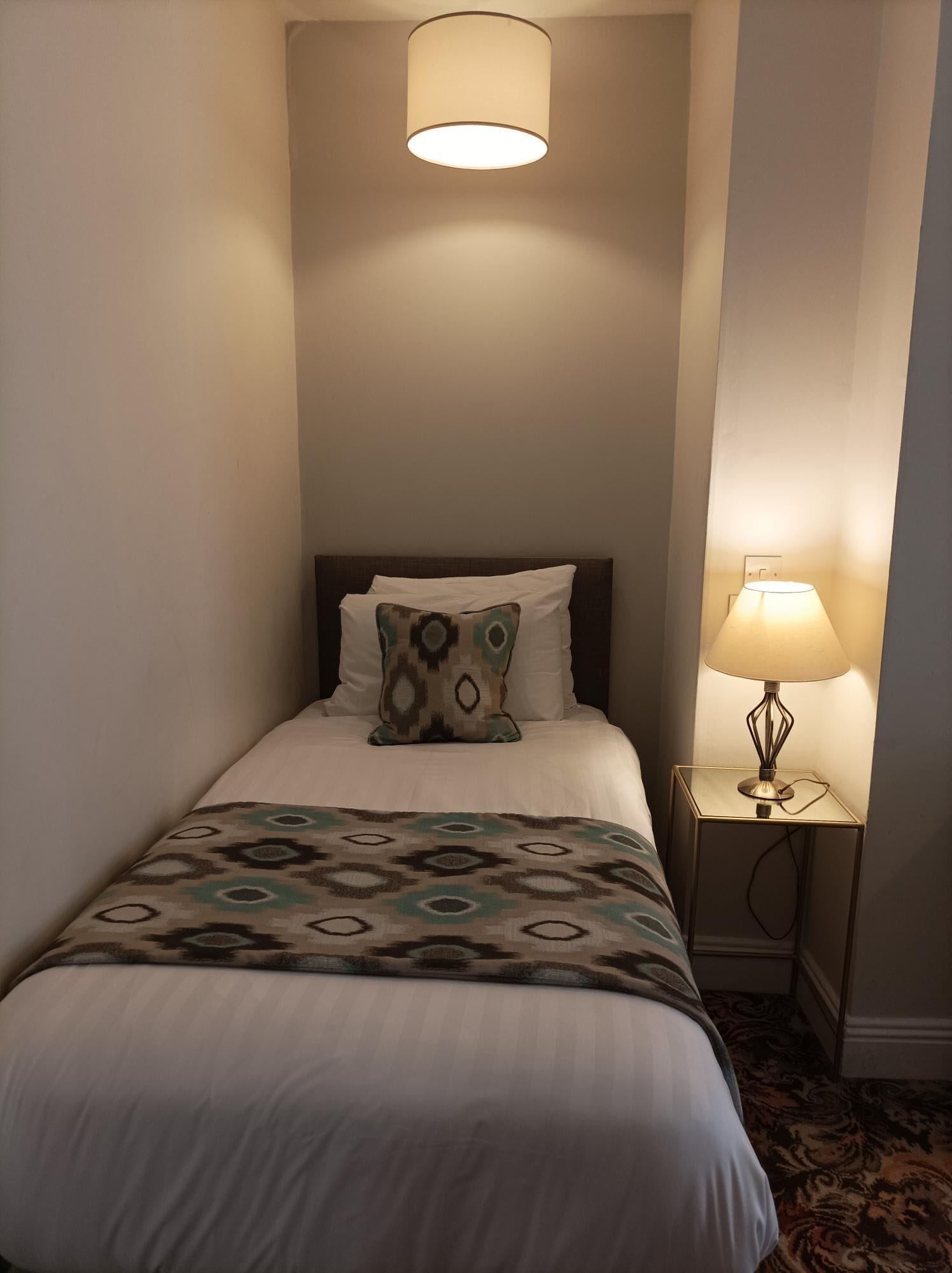 Uma cama bem arrumada com um cobertor estampado, um travesseiro decorativo e uma luminária de cabeceira.