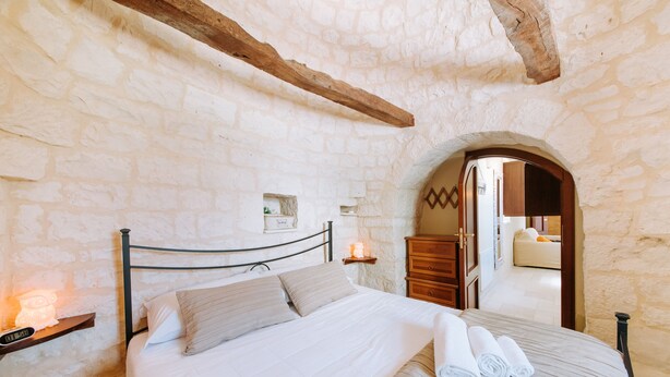 Un dormitorio con pared de piedra, viga de techo de madera, cama con ropa blanca, mesita de noche con lámpara y cómoda de madera.