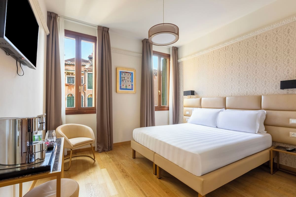 A hotel room with a bed, a desk with a coffee maker, a chair, a TV, and a window with curtains.