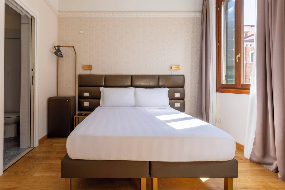 A hotel room with a large bed, a bedside table, a lamp, and a window with curtains.