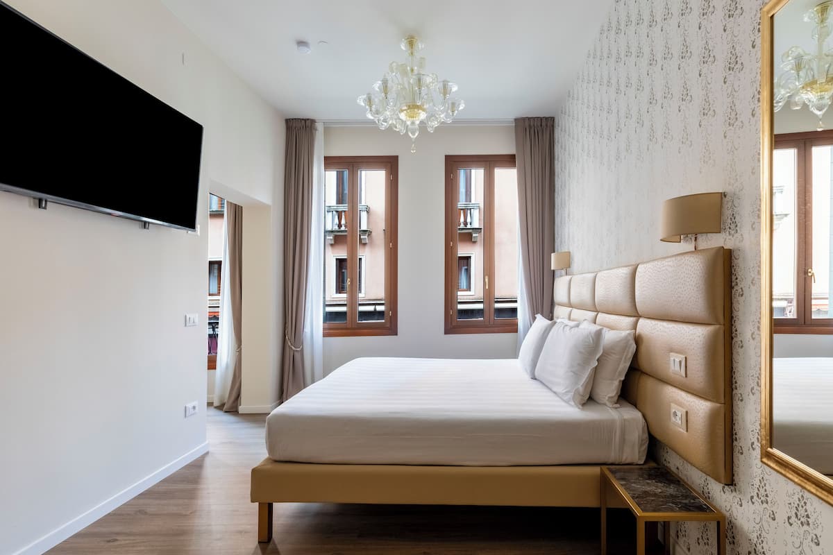 A hotel room with a large bed, a flat-screen TV, a chandelier, and a window with curtains.