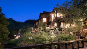 Un bâtiment japonais traditionnel, à plusieurs niveaux et doté d’une façade en bois, entouré d’une végétation luxuriante et traversé par un petit ruisseau.