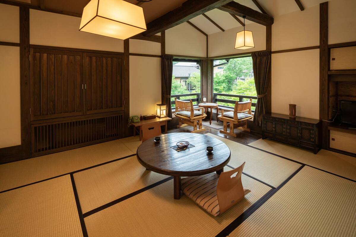 A traditional Japanese-style room with tatami flooring, a round table with cushions, wooden furniture, and a view through large windows.